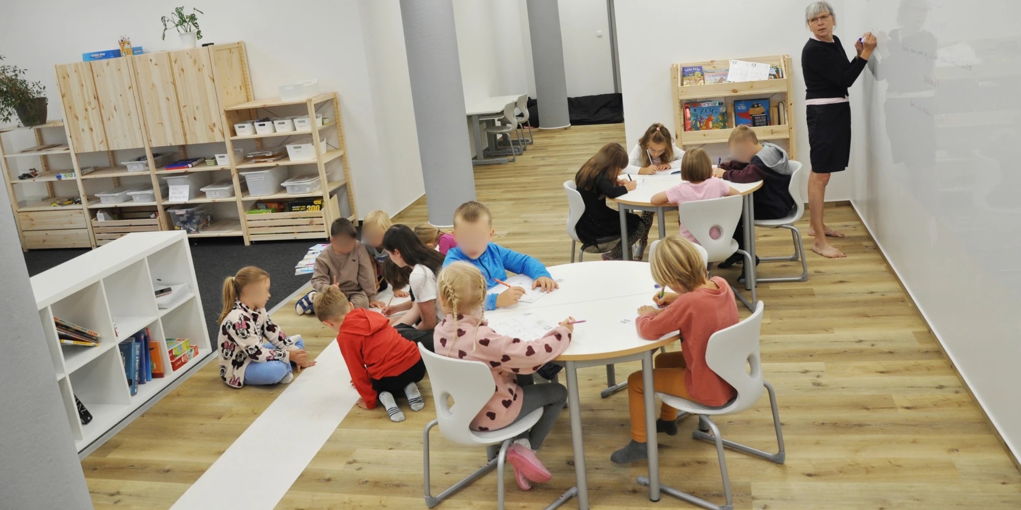 Třída při práci – děti píšou u kulatých stolů i vsedě na koberci, pedagožka píše na tabuli